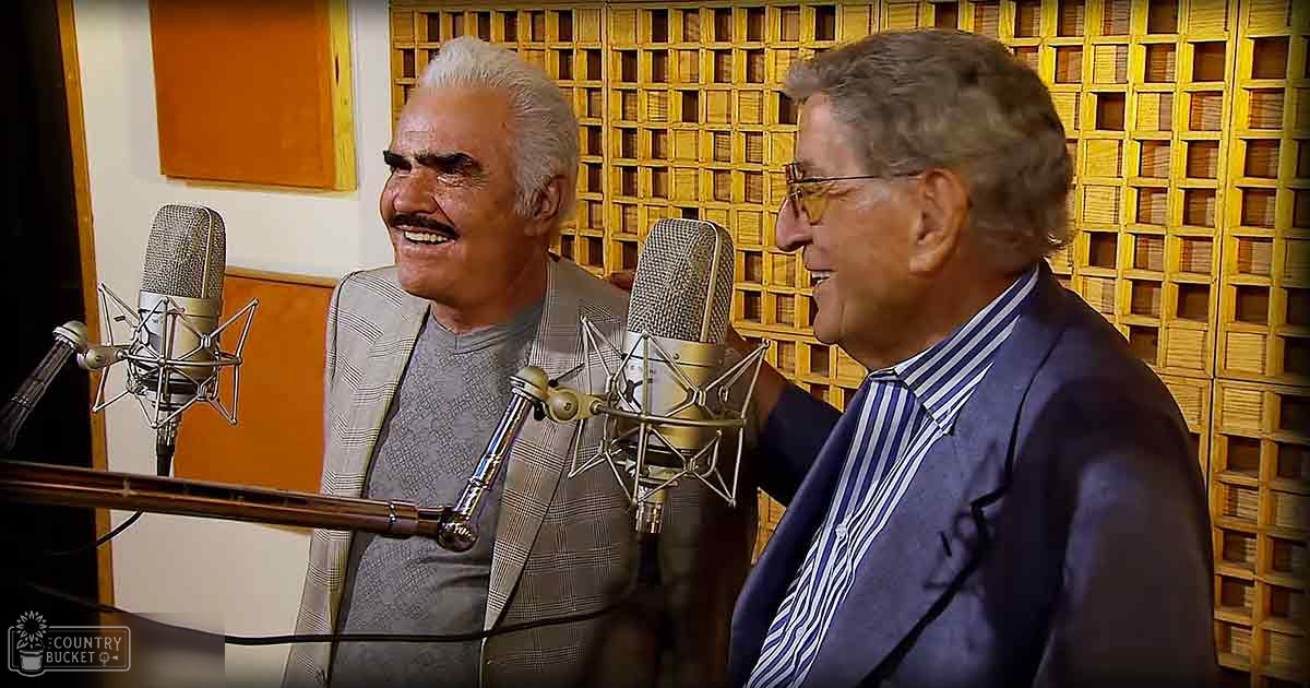 Middle-aged to older adult male in a gray blazer with checkered pattern, recording studio setting with soundproofing panels and two microphones.