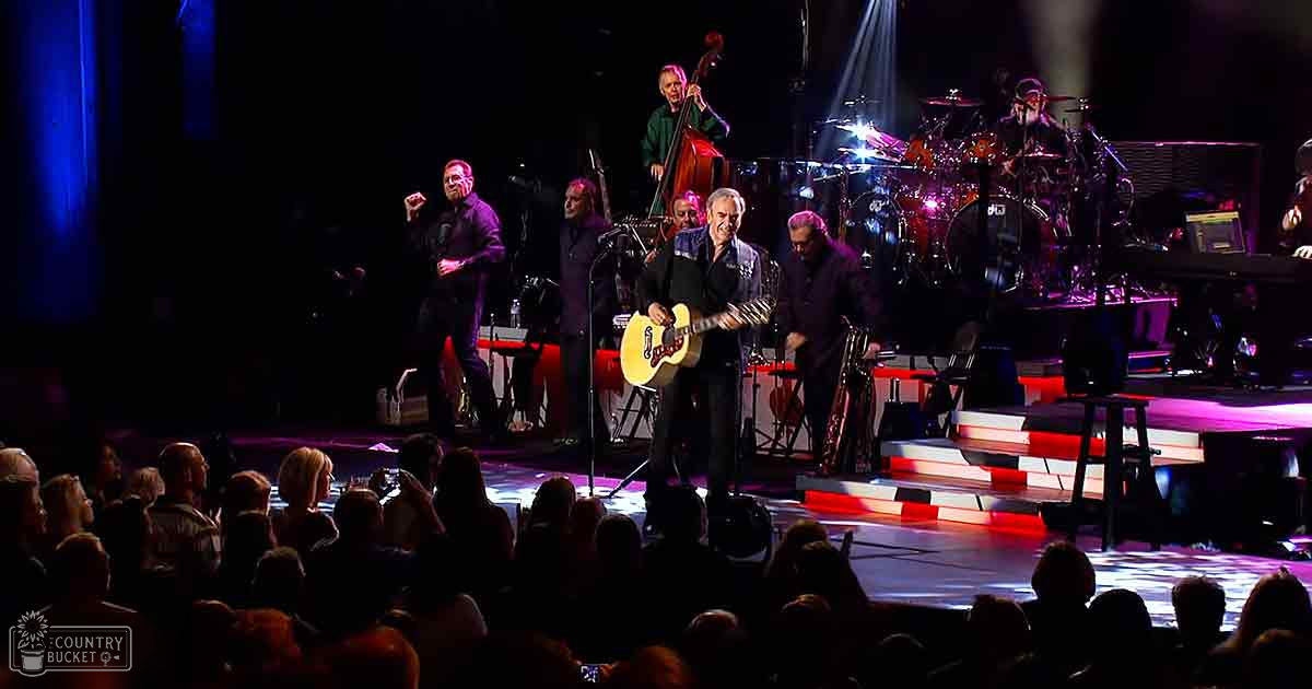 Indoor concert with musicians playing guitars, microphones present, dark curtains backdrop, audience seated, artificial stage lighting, dark clothing, red and white steps visible.