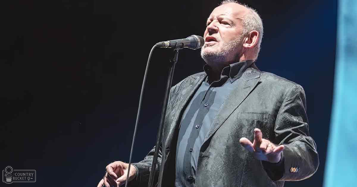 A middle-aged male singer in a relaxed fit, dark gray blazer with subtle pattern and lapels, performing on stage at an event with 'Madly Odd' branding visible.