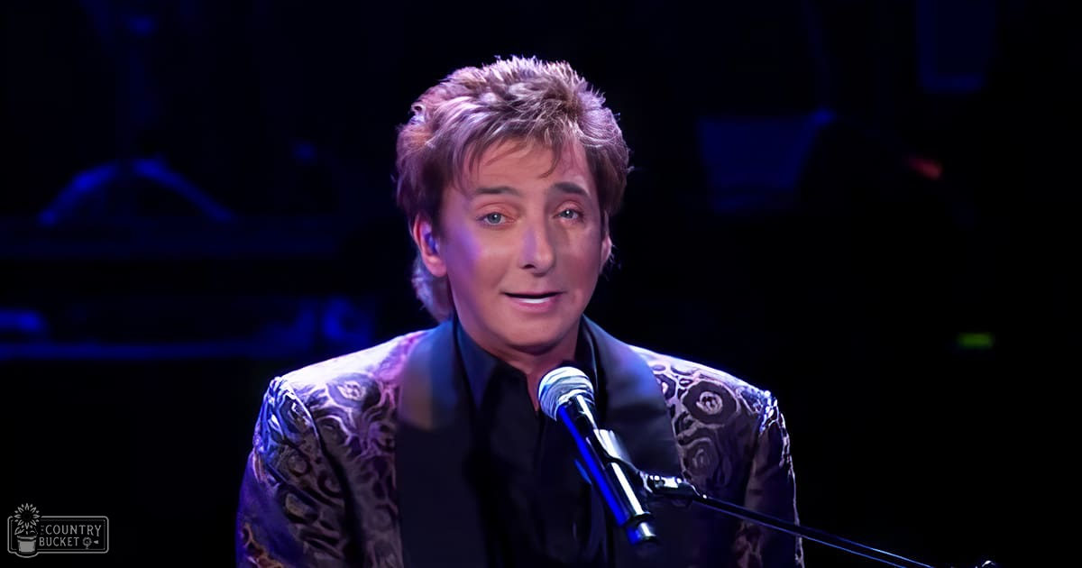 A middle-aged male performer wearing a patterned blazer over a black shirt and tie stands on stage with a microphone stand, illuminated by blue stage lights.