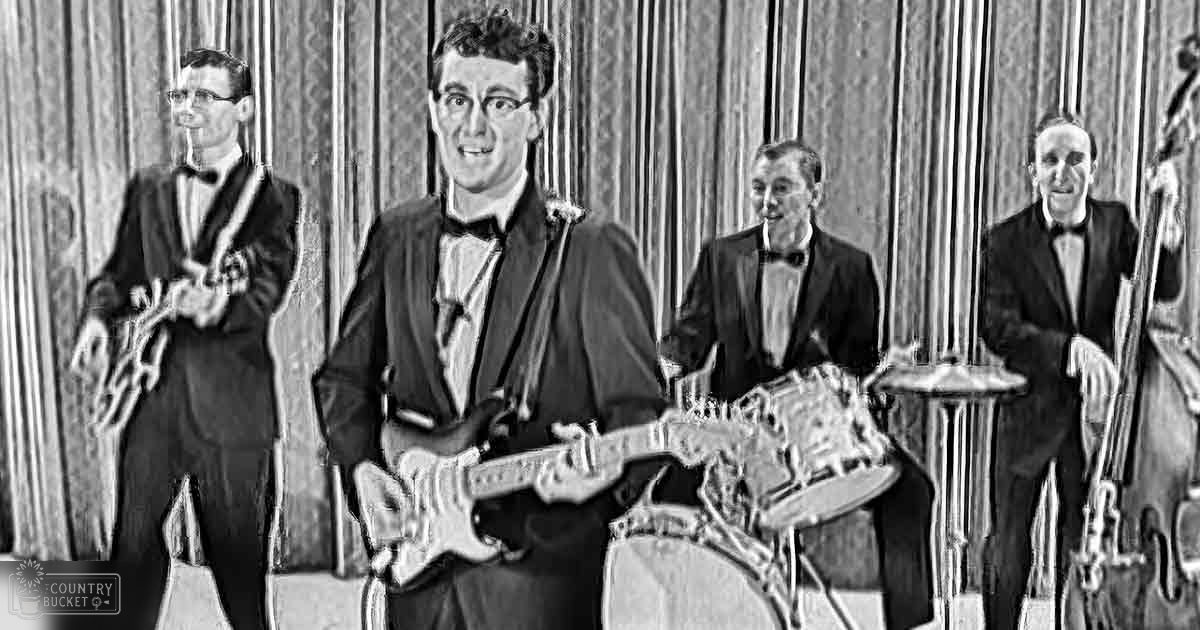 A young adult male singer in glasses plays electric guitar, surrounded by other musicians playing various instruments under artificial indoor lighting.
