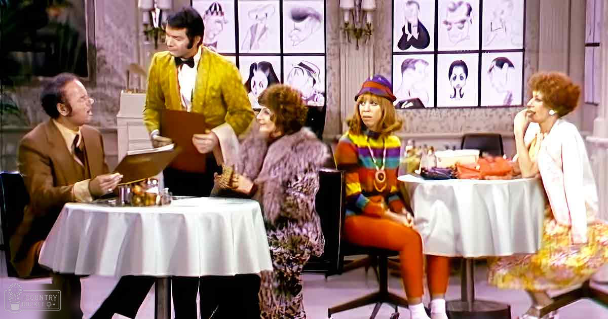 A group of individuals in a brightly lit indoor setting resembling a television studio with various furniture and decor items, including round tables covered with white tablecloths and framed sketches on the wall.