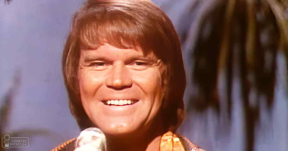 An adult male wearing a textured, patterned jacket with V-neck style and short sleeves, likely from the 1970s, indoors with vertical lines suggesting curtains or blinds in the background.