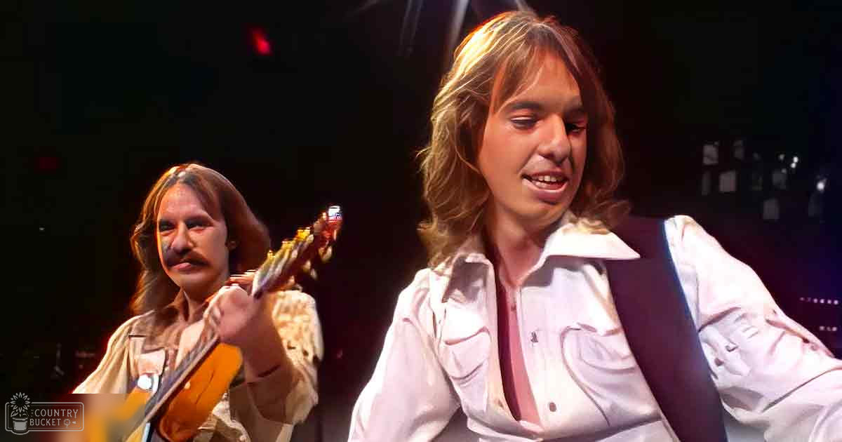 Young adult male musician performing on stage with another individual playing guitar in the background under dim lighting.