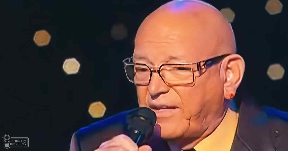 An older adult male, possibly middle-aged, in a dark suit jacket with light-colored shirt and rectangular-framed glasses, holding a microphone on stage under artificial lighting.