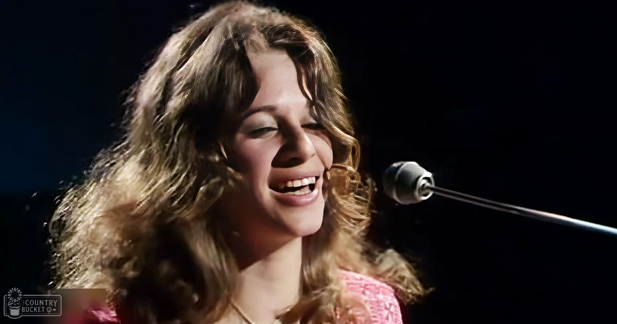 A young adult female singer wearing a pink lace-trimmed top performs into a microphone in an indoor performance setting with dramatic lighting.