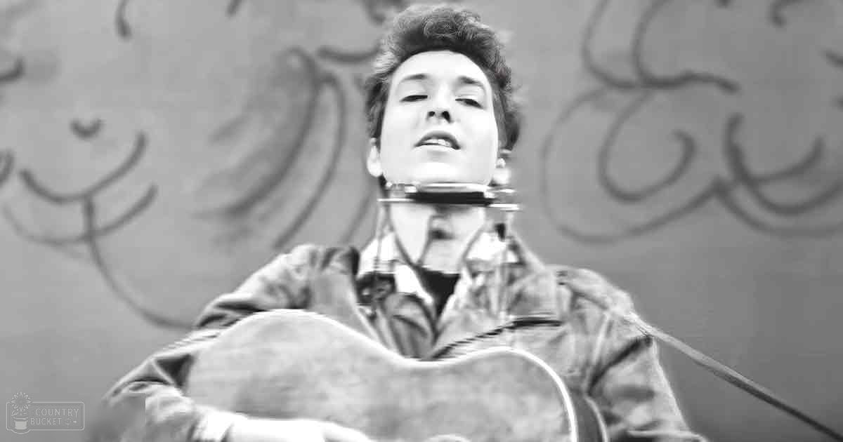 A young adult male plays an acoustic guitar against a graffiti-covered wall in an urban setting under natural daylight.