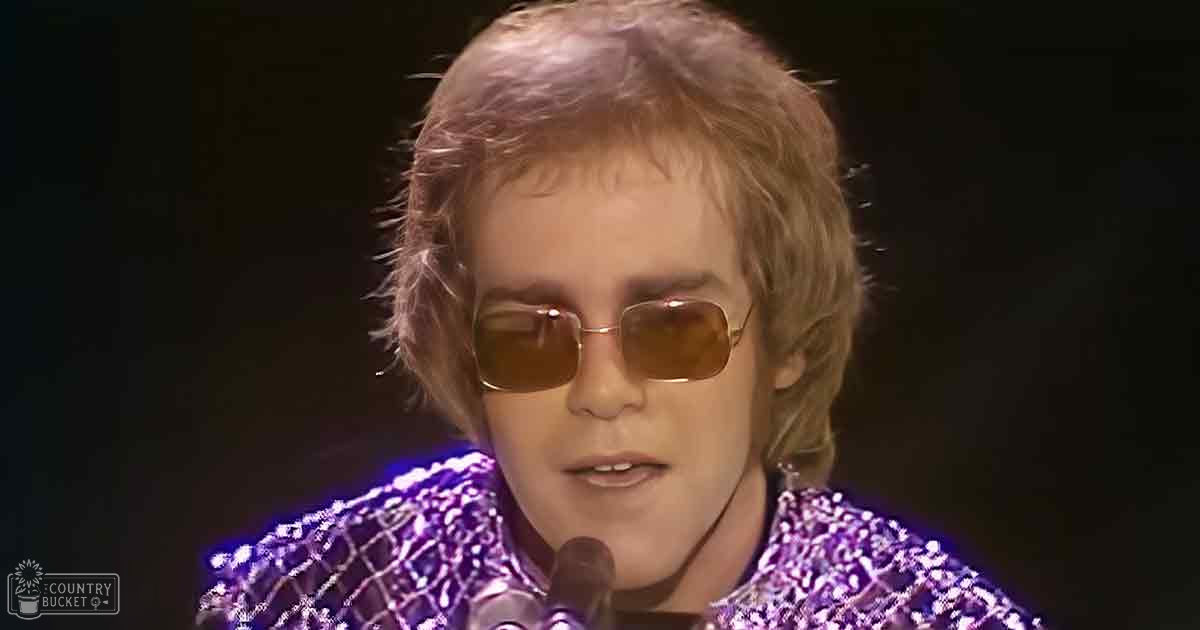 A young adult male singer in a stage costume with intricate patterns and sequins, performing on an indoor stage under dark lighting.