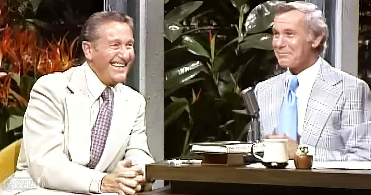 Two older adults in a studio setting, wearing formal attire with beige suit jackets and patterned blazers. A dark wooden table holds a mug, papers, and pens, alongside a microphone suggesting an interview or talk show format.