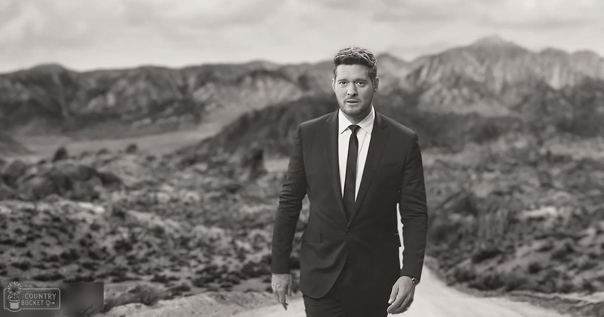 An adult male wearing a dark suit jacket and trousers stands outdoors with rugged mountains in the background under an overcast sky.