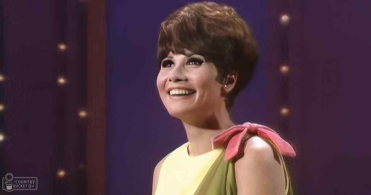 Adult female wearing a sleeveless yellow dress with pink accent and bow detail, possibly mid-20th century stage attire, indoors on what appears to be a stage or studio set.