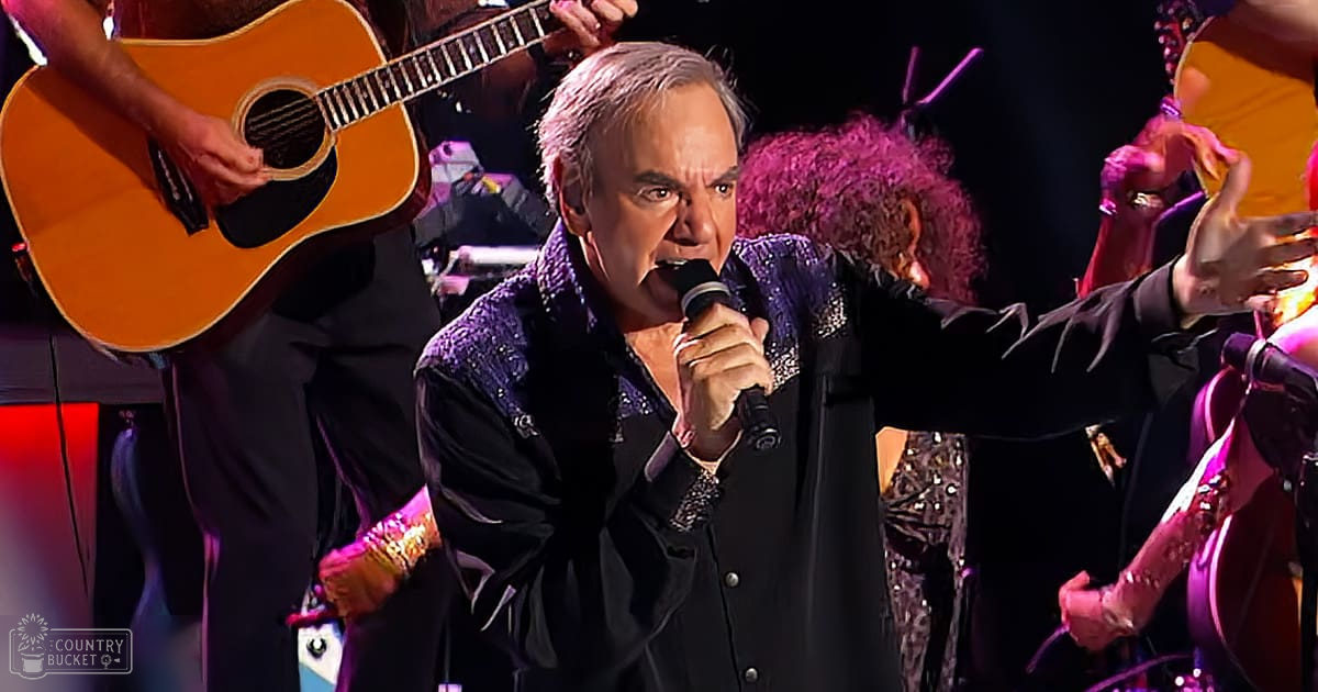 A middle-aged male singer performs on stage at an indoor concert venue, surrounded by musicians wearing black attire with embellishments and rings.