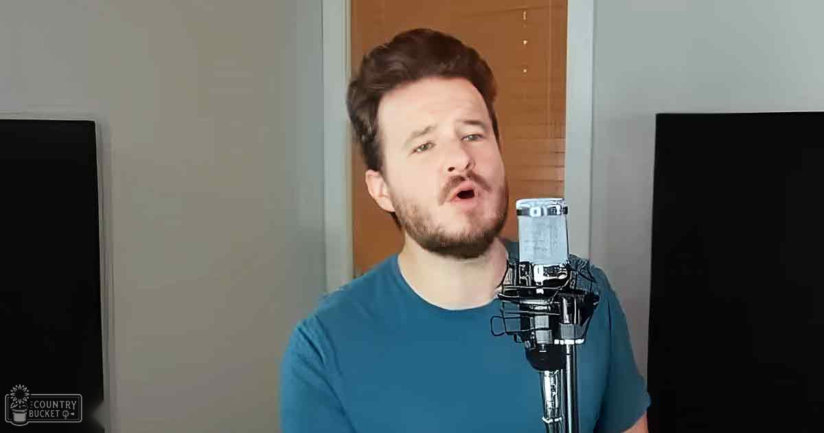 A young adult male wearing a teal, solid-color t-shirt with short sleeves and a round neckline stands indoors against plain walls, with a microphone setup in front of him.