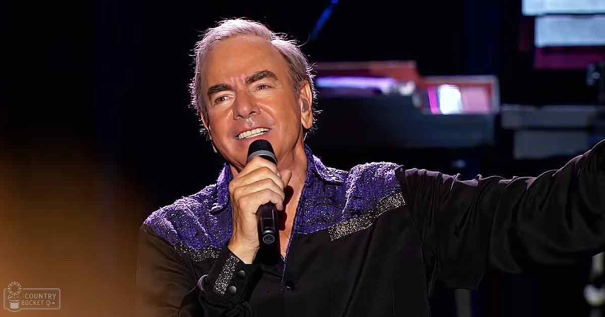A middle-aged adult male singer in a relaxed, shiny black shirt with purple accents on stage at an indoor concert venue.