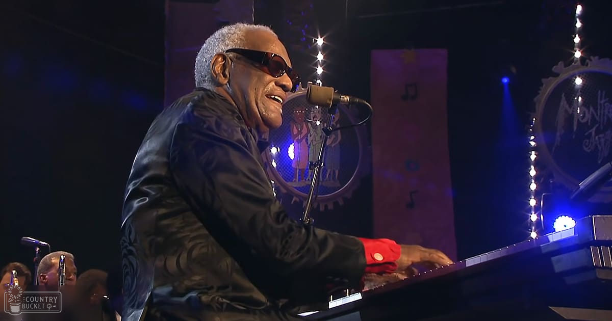 A middle-aged man with gray hair wearing sunglasses and a black leather jacket plays piano and sings into a microphone on an indoor stage at Montreux Jazz Festival.