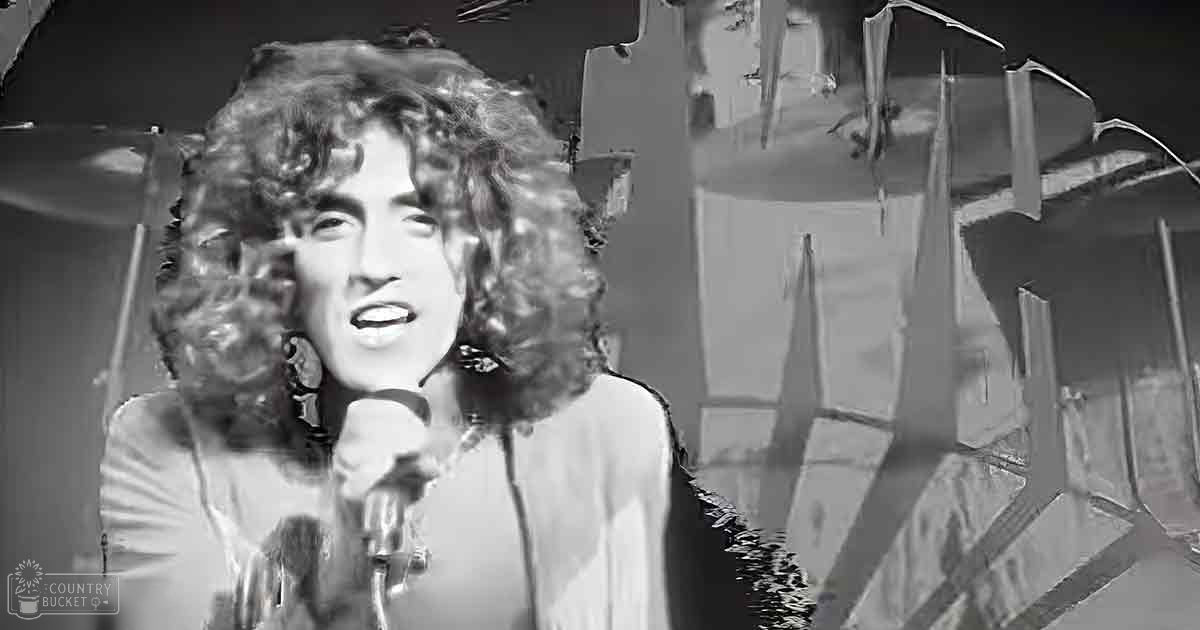 An adult female performer in a textured, light-colored top with long sleeves, holding a microphone on stage against a patterned backdrop.