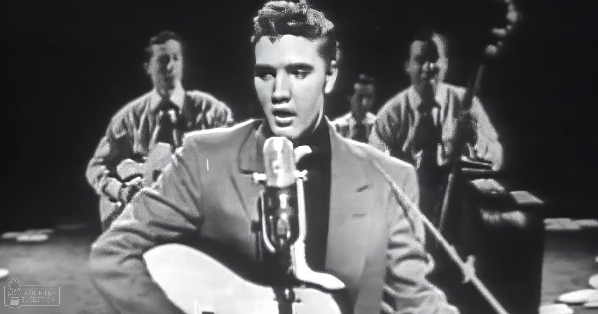 Adult male singer performing on stage with a light-colored suit and dark tie, microphone stand in foreground; young adult playing drums in background at night.