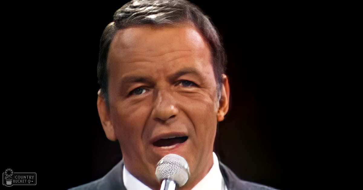 A middle-aged adult male in a formal suit stands on stage, wearing a dark gray or black tailored suit with a white dress shirt, under controlled indoor lighting.