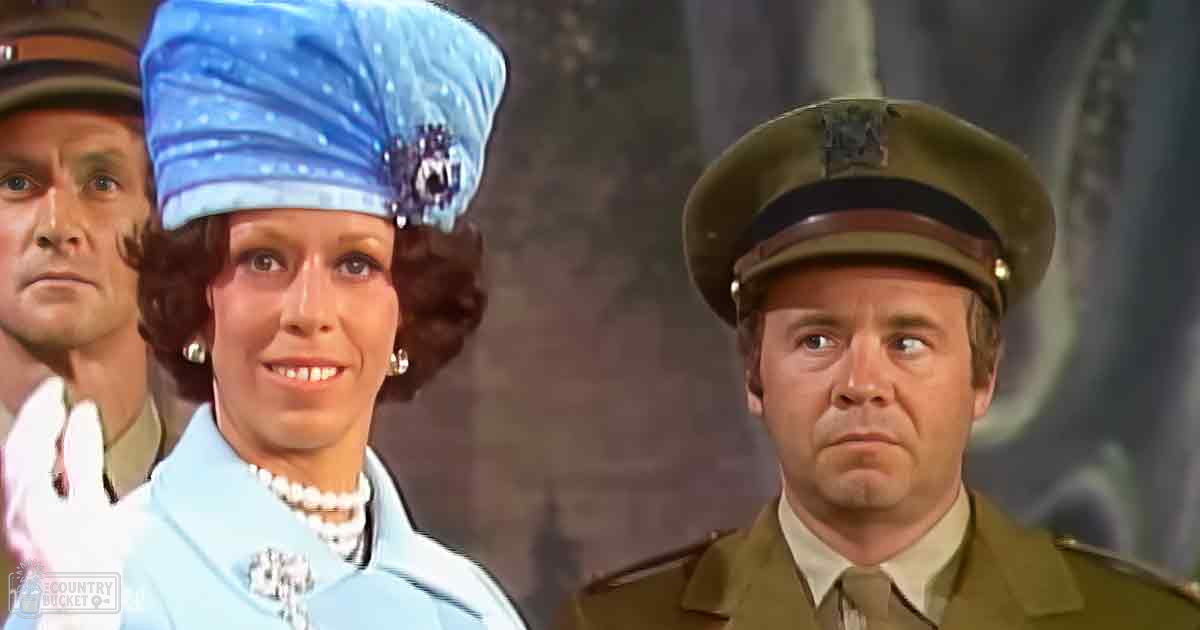 Adult female wearing a light blue coat, brooch, pearl necklace, large earrings, and headscarf with decorative element, accompanied by two men in military uniforms indoors.