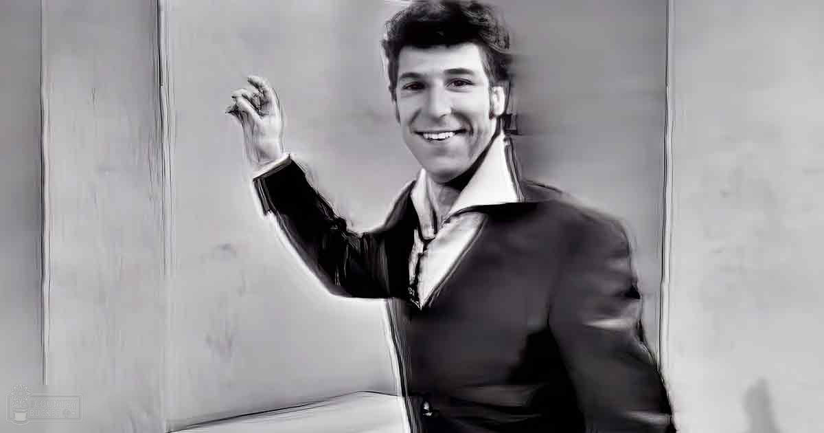 A young adult male wearing a dark blazer over a light shirt indoors with natural lighting.