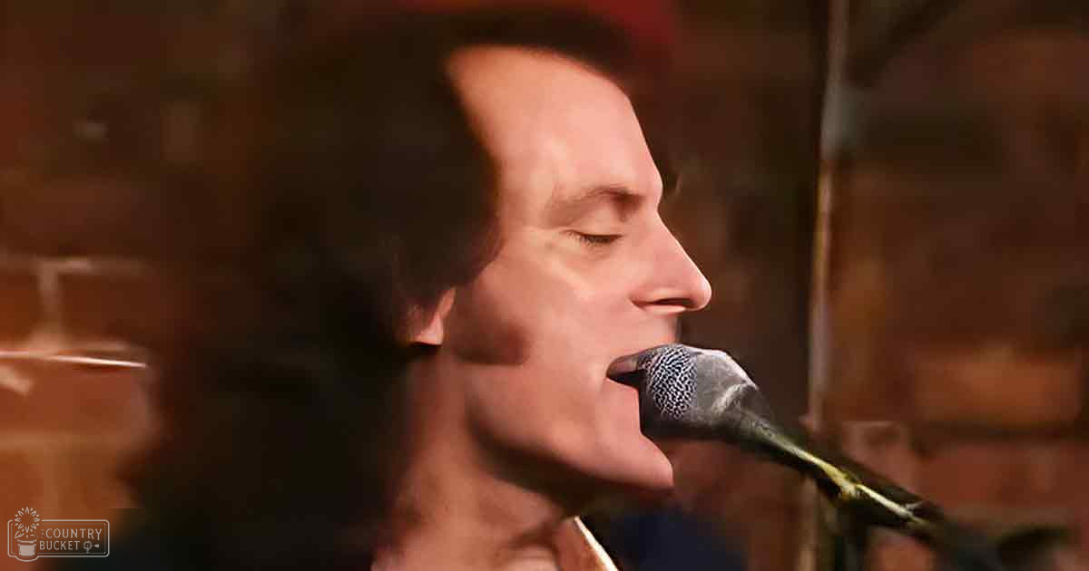 A middle-aged man singing into a microphone in an indoor setting with brick walls, dimly lit environment suggesting evening or night time.