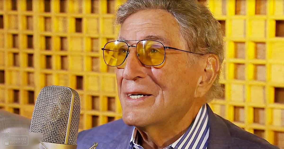 An older adult male in a recording studio, wearing a dark blazer over a striped shirt with vertical blue and white stripes, standing near a microphone against yellow acoustic foam panels.