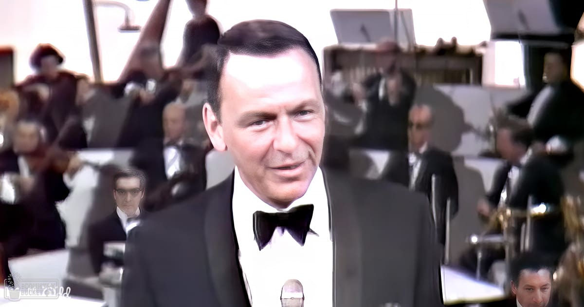 Adult male in a black tuxedo with a white shirt and black bow tie standing on stage, holding a microphone. Background shows an orchestra playing various musical instruments under well-lit stage lights.