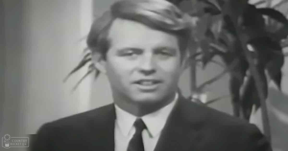 A young adult male in a dark suit jacket, white dress shirt, and solid-colored tie stands indoors with indoor plants in the background.