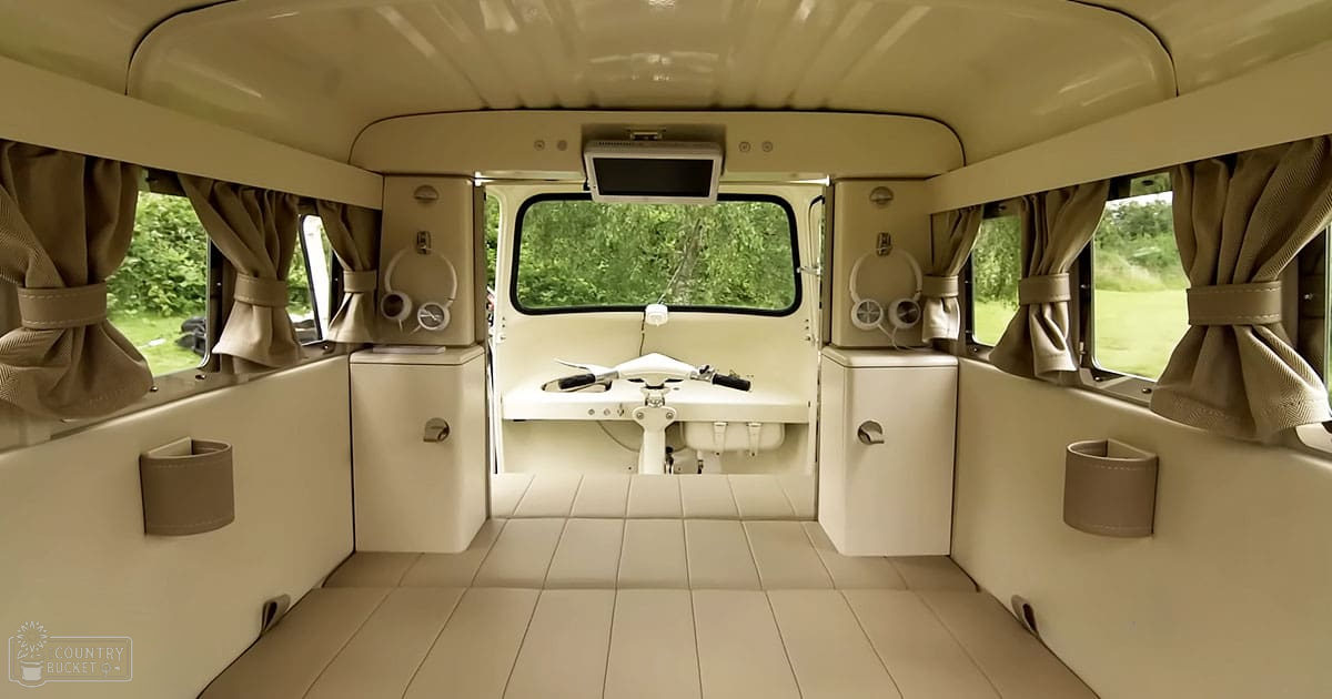 Van interior setup as a camper or mobile home, small sink, toilet with seat cover, headphones hanging from hooks, tied-back curtains allowing natural light, beige and cream walls, brown curtains, daytime outdoor greenery.