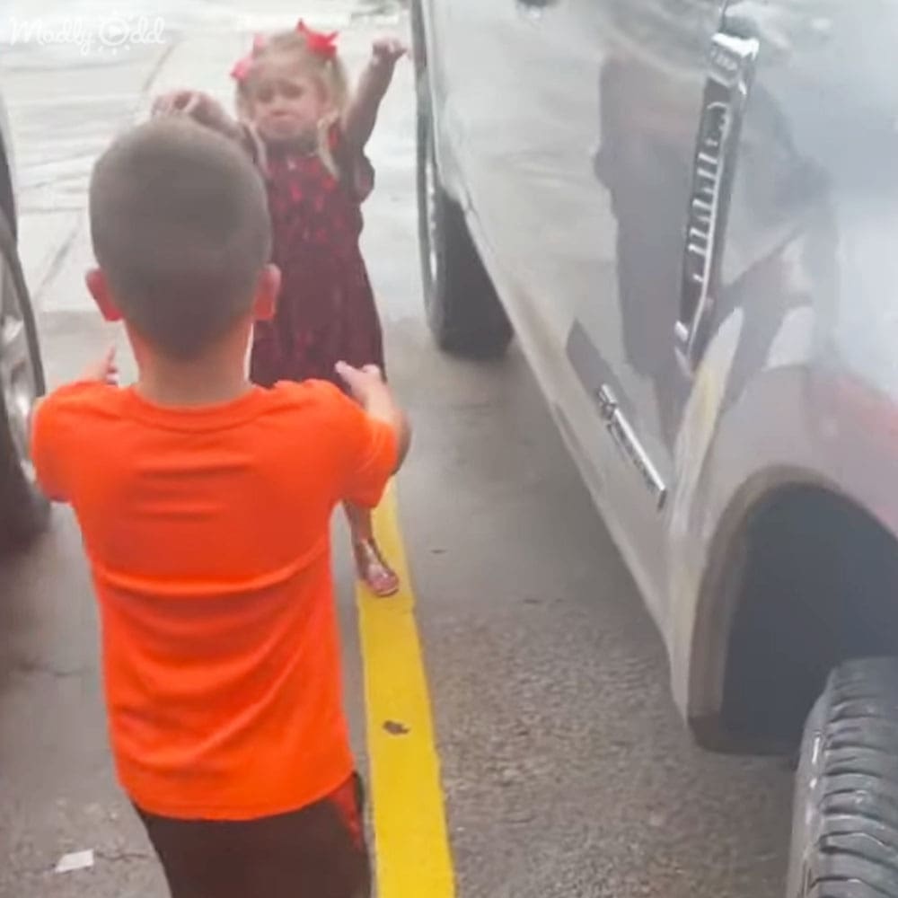 A toddler girl wearing a dark red floral dress and red bows stands near an orange-shirted toddler boy in an outdoor parking area with vehicles visible, suggesting spring or summer.