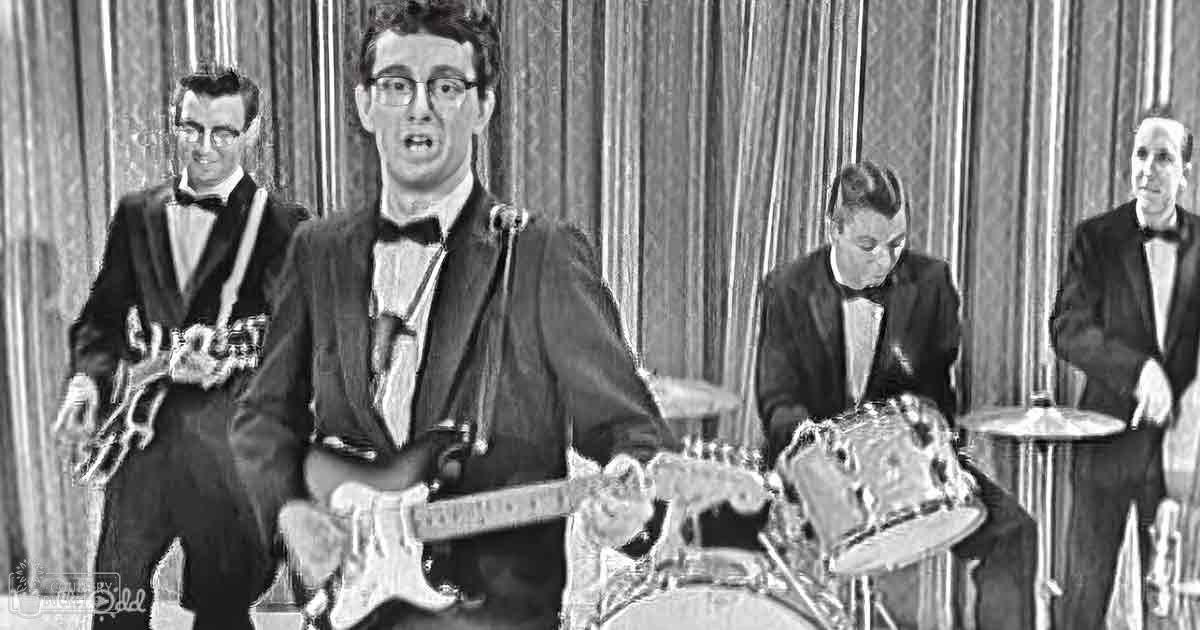 A young adult male plays an electric guitar in a brightly lit indoor studio setting with other band members visible. Musical instruments and curtains are present, suggesting a recording or performance environment.