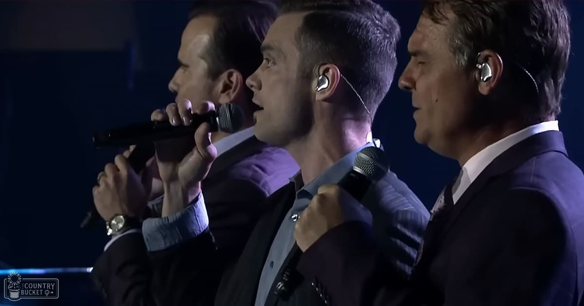 Three adult male singers performing on stage at an indoor concert, wearing blazers and button-up shirts under stage lighting.
