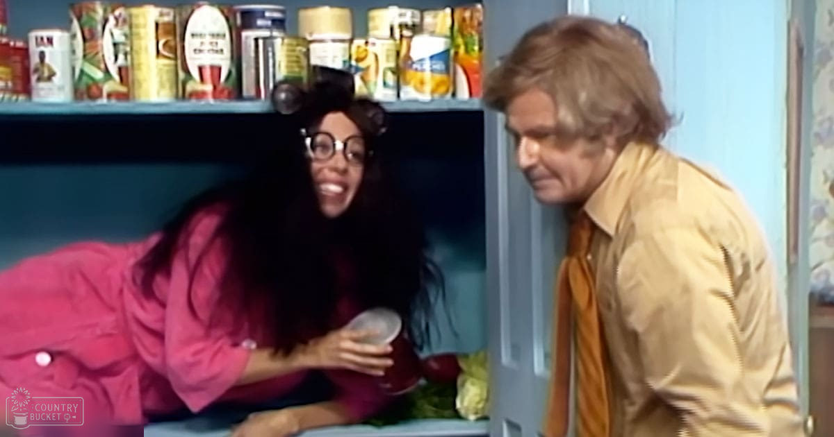 A woman wearing a pink 1970s ruffled shirt and glasses stands in a kitchen, holding a clear plastic container; an adult male in a beige jacket with yellow shirt and orange tie is nearby. The setting includes shelves of canned goods and floral-patterned wallpaper.