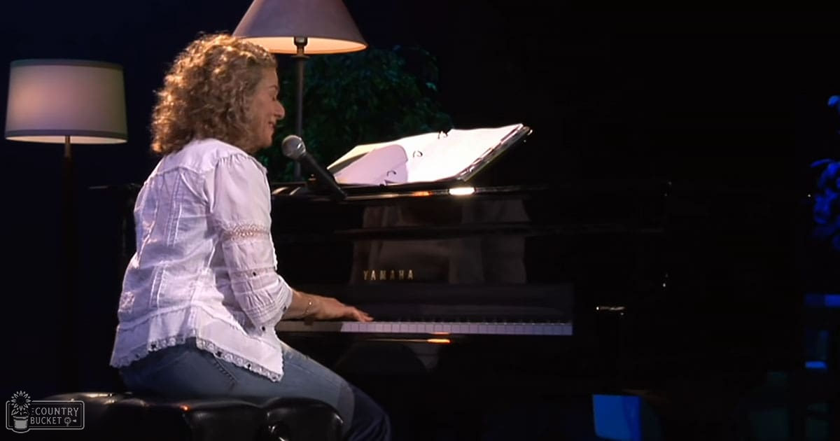 A middle-aged woman in a white ruffled blouse and blue jeans plays a Yamaha grand piano indoors under artificial evening lighting with two floor lamps nearby.