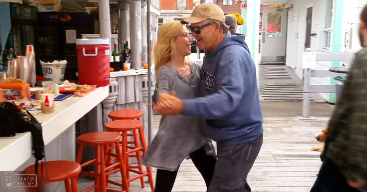 A middle-aged couple dancing outdoors on a boardwalk with a small dog lying nearby and food stands in the background.
