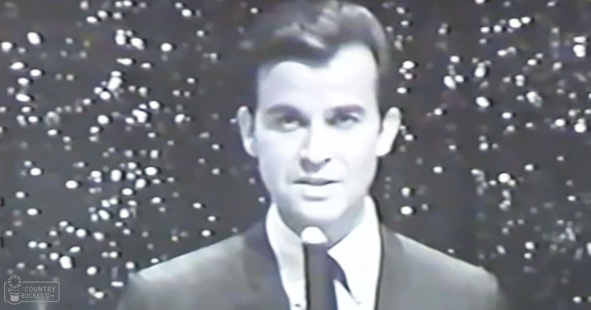 A young adult male wearing a classic dark suit jacket, white shirt, and dark tie stands indoors with a backdrop of small dots.