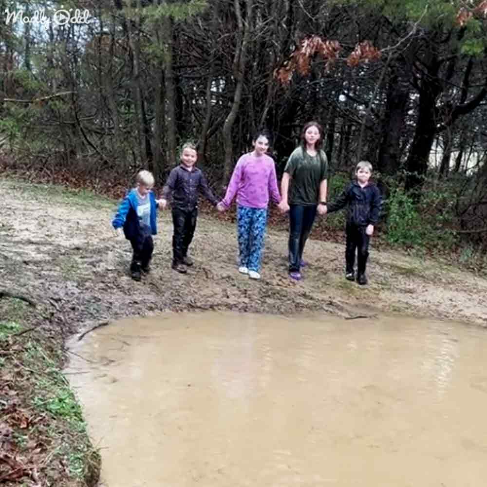 A group of children and an adult female exploring a wooded area near a muddy puddle, late autumn or early spring with bare trees and green foliage.