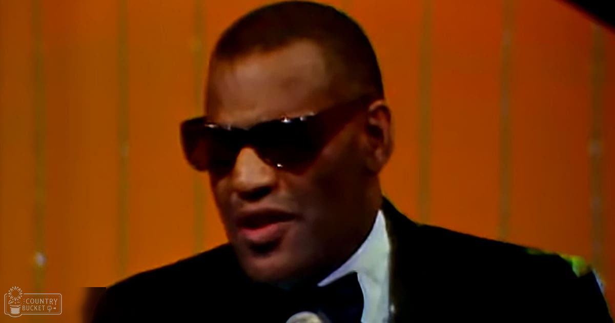 Adult male wearing a black mid-century to early 21st century suit with white shirt and dark bow tie, standing against an orange striped background in daytime lighting.