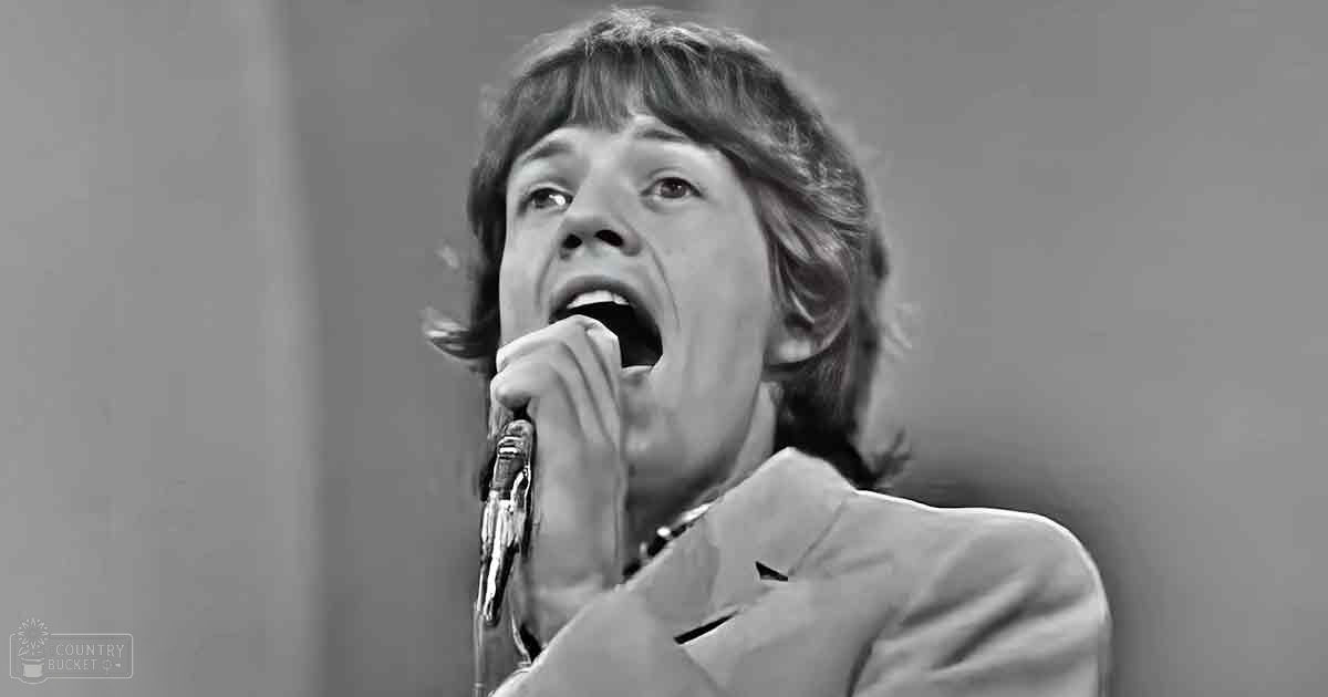 A young adult male in a mid-20th century-style suit jacket and shirt performs on stage under dim lighting, holding a microphone.