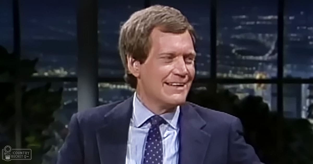 Middle-aged man in a dark blue suit jacket with white polka dot tie and light blue dress shirt, set against an evening cityscape studio backdrop.
