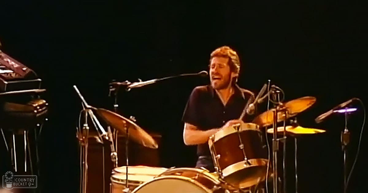 A middle-aged male drummer performing on stage with a dark-colored, casual button-up shirt in an indoor concert setting.