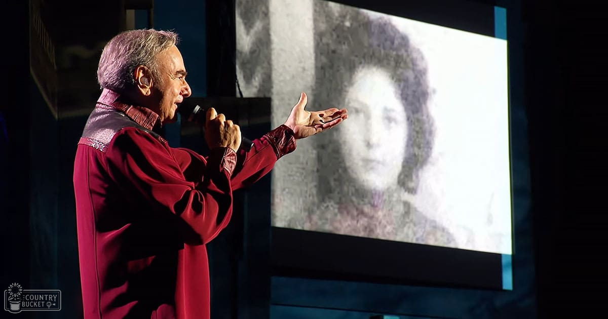A middle-aged man in a red and purple silk or satin robe giving a presentation at an indoor conference or seminar with stage lighting focused on him, surrounded by dark walls and a large projection screen.