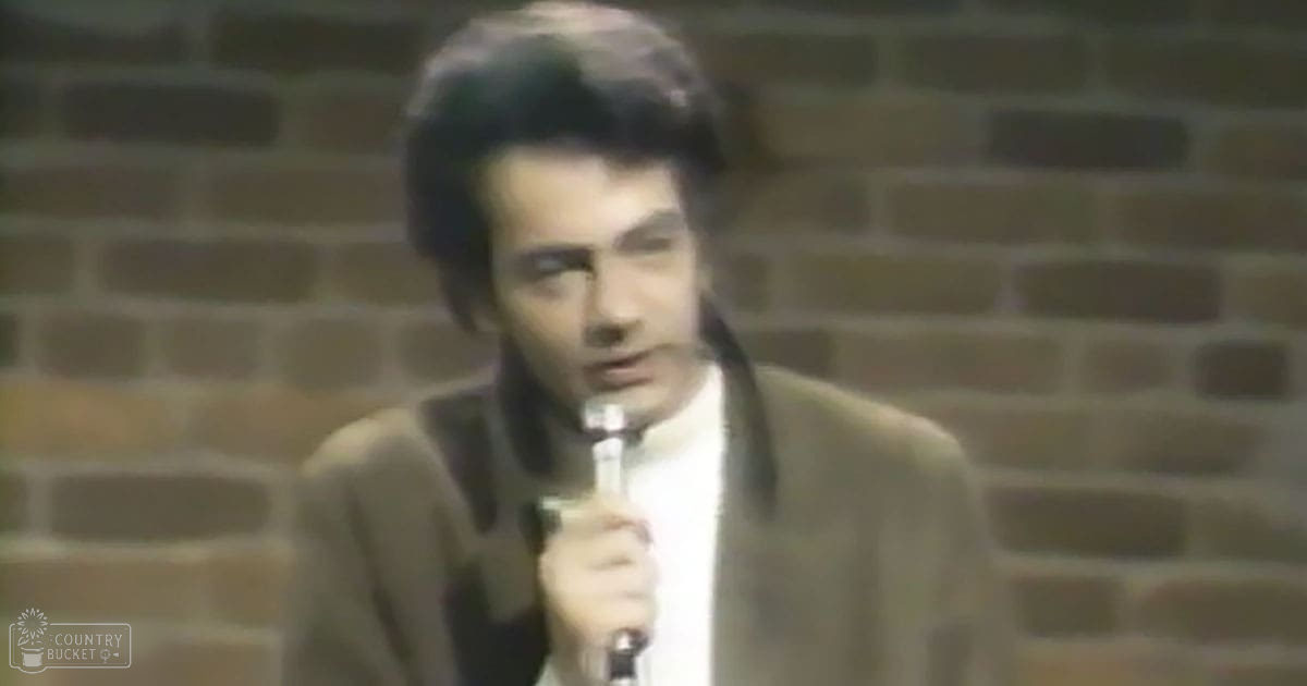 A young adult male in a brown suit jacket with 'Madly Odd' printed on the bottom edge, performing on stage against a brick wall under even and bright lighting.