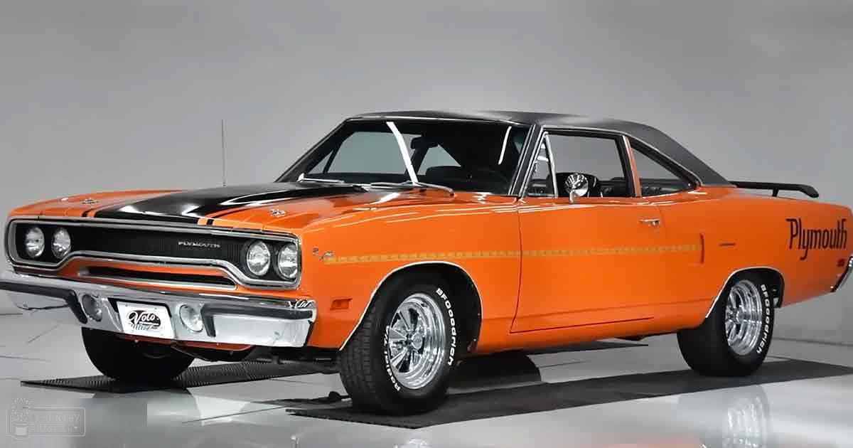 A classic Plymouth car with bright orange body, black roof, white racing stripes, and chrome details displayed in an indoor showroom setting.