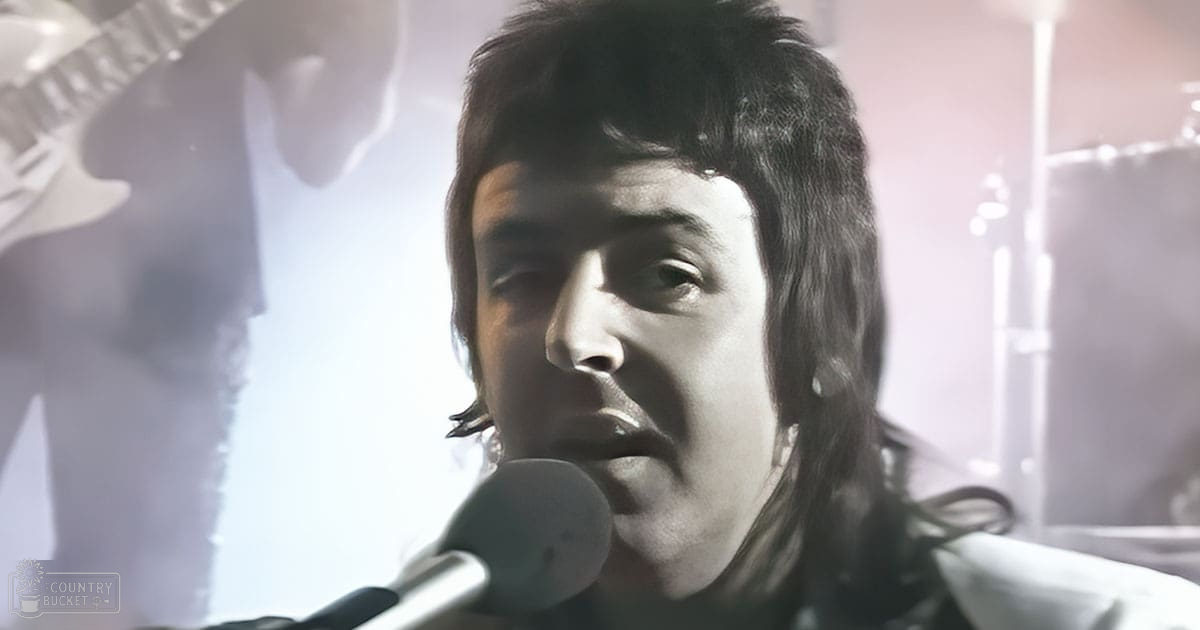 An adult male singer performing at an indoor concert venue with stage lighting and musical instruments in view.