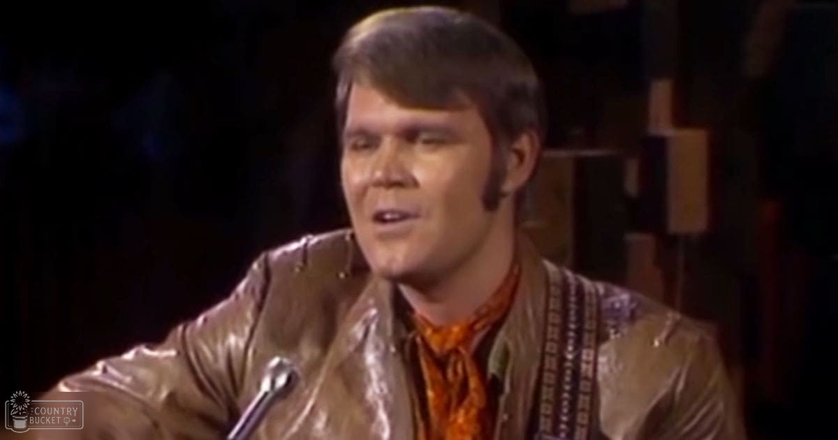 A middle-aged adult male in a relaxed, slightly loose-fitting brown leather jacket with an orange and patterned fabric underneath stands indoors near a microphone stand and wooden structure.