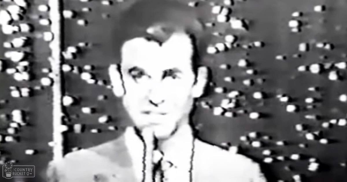A young adult male wearing a white shirt with a collar and what seems like a dark tie or scarf, set against an indoor industrial backdrop.