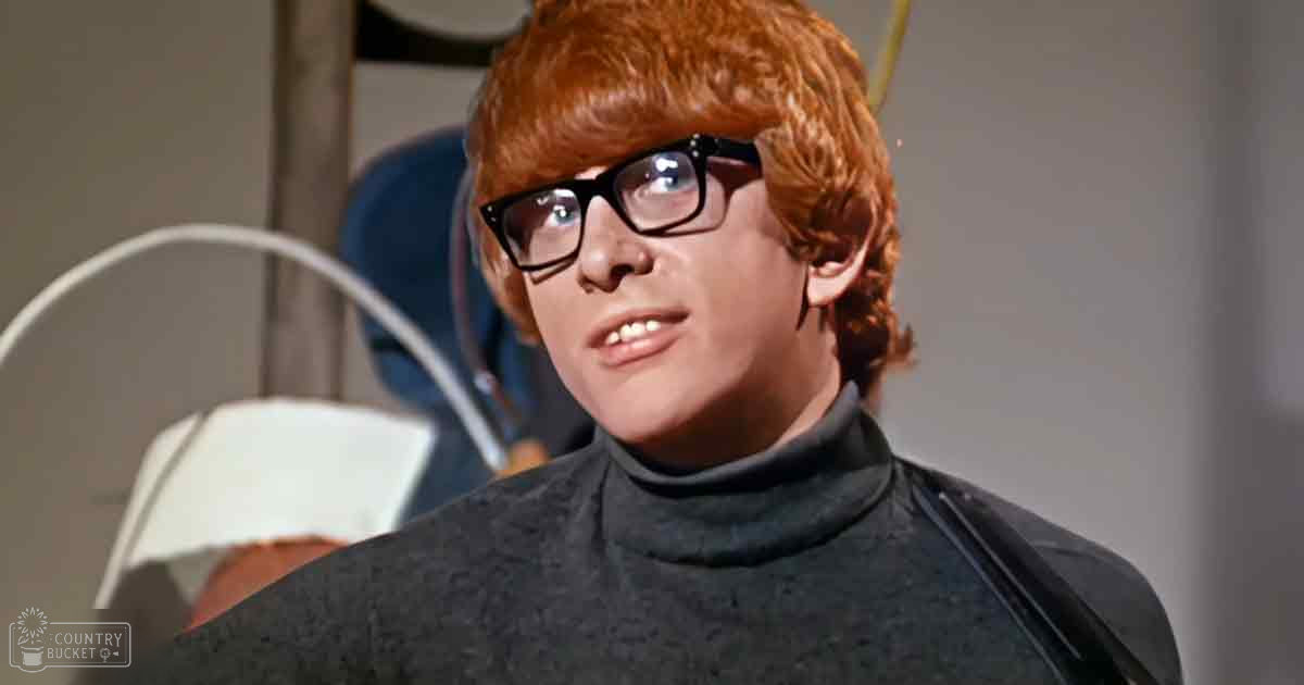 Young adult male wearing a dark gray/black turtleneck sweater with high neckline and long sleeves, indoors in an even-lit space suggesting artificial light.