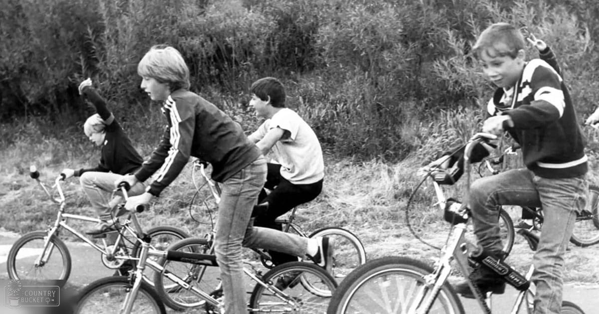 Group of children riding bicycles outdoors, wearing dark jackets with white stripes and light-colored pants; setting appears to be a grassy area with bushes, likely late spring or summer.