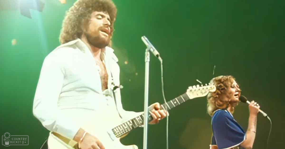 Adult male musician wearing a white open-collared shirt with cotton-like material and an adult female performer dressed in a blue sleeveless top during a live concert setting, stage lighting present.